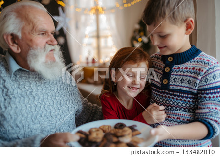 Kids with senior grandfather indoors at home at Christmas, eating biscuits. Kids with senior grandfather indoors at home at Christmas, eating biscuits. 133482010