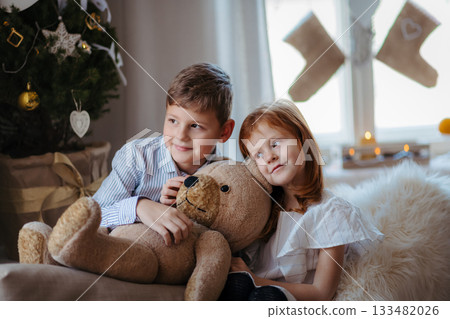Small girl and boy indoors at home at Christmas, holding teddy bear. 133482026
