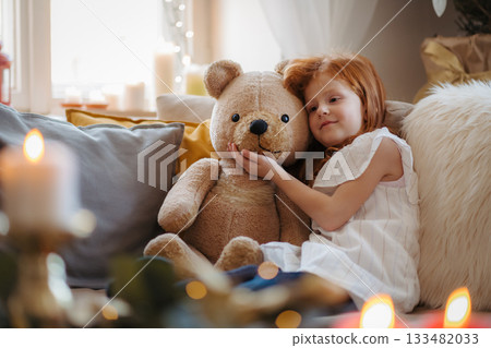 Small girl sitting on sofa indoors at home at Christmas, hugging teddy bear. 133482033