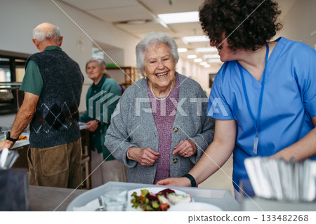 Senior woman having lunch with supportive caregiver in community center cafeteria. Senior woman having lunch with supportive caregiver in community center cafeteria. 133482268