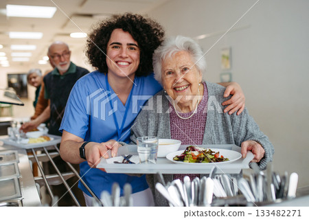 Senior woman having lunch with supportive caregiver in community center cafeteria. 133482271