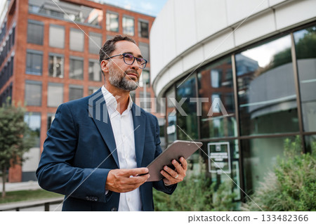 Businessman in front of office building, holding tablet and reading something. 133482366
