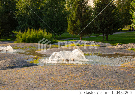modern water fountain in the garden, lush greenery and stone pathways. The warm light of sunset enhances the calm ambiance of the area. 133483266