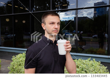 Young Caucasian man enjoys coffee outdoors, sipping coffee from disposable white cup. The scene captures bright day with reflections in the glass surfaces around him, showcasing urban life 133483445