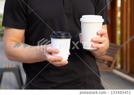 Man holds two coffee cups in outdoor setting during sunny afternoon in casual environment 133483451