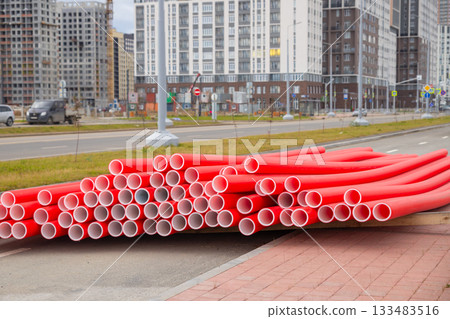 Stack of bright red new plastic pipes on the street. Modern buildings rise in the background, urban development Stack of bright red new plastic pipes on the street. Modern buildings rise in the background, urban development 133483516