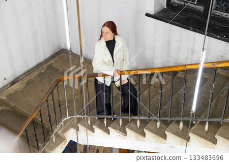 A Caucasian woman thoughtfully walks up the stairs with a phone in her hands. doctor, mother of two children, professor, businesswoman with long red hair 133483696