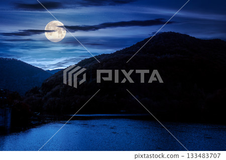 mountainous landscape with lake in autumn at night. forest on hills near the shore of river under dark sky in full moon light. beautiful view of stunning place 133483707