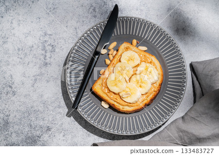 Peanut butter toast and banana on a dark plate on a gray background with knife Peanut butter toast and banana on a dark plate on a gray background with knife 133483727