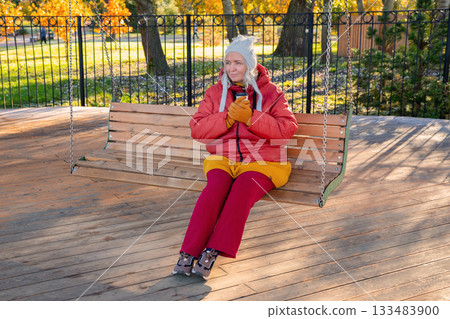 A body-positive Caucasian woman with a disposable cup of coffee sits on a wooden swing in an autumn park in the morning. The mother of two is relaxing alone. Mental health, harmony with oneself, 133483900