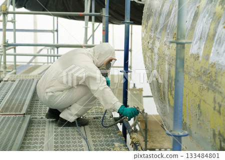 Worker in protective gear sprays paint on large metal tank while crouching on scaffolding at industrial site during daylight hours 133484801