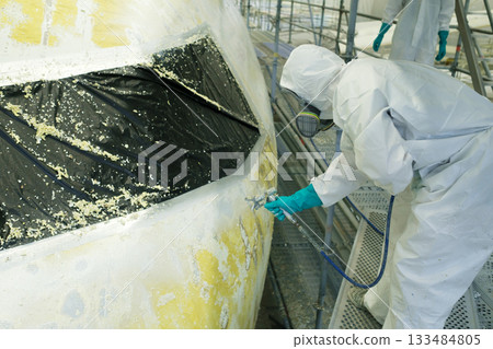 Professional worker applies spray paint to an aircraft in a hangar during repairs, ensuring precision and safety while wearing protective gear Professional worker applies spray paint to an aircraft in a hangar during repairs, ensuring precision and safety while wearing protective gear 133484805