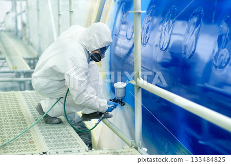 Worker sprays paint on aircraft fuselage in industrial facility during daytime under bright lights to ensure precision and quality in finishing 133484825