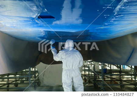 Worker applies blue paint to the underside of a boat in a well-lit workshop during daytime, ensuring precise and even coverage for protective finishing 133484827