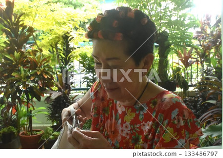 Asian elder woman sewing cloth for repairing in home front yard 133486797
