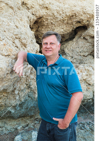Portrait of an adult man in a blue shirt standing near a rock on the beach 133488937