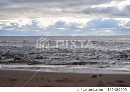 Waves on sandy shore with stones in foreground under cloudy sky over Gulf of Finland. Waves on sandy shore with stones in foreground under cloudy sky over Gulf of Finland. 133489803