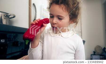 Little girl examining and eating red pepper in home kitchen Little girl examining and eating red pepper in home kitchen 133489942