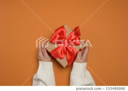 Two female hands in a white knitted sweater hold a square gift box wrapped in craft paper with a large red satin bow. 133490990