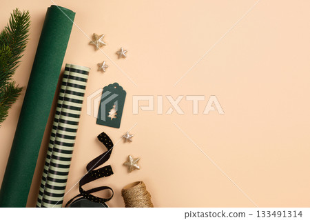 Christmas wrapping paper rolls, green gift tag, and gold stars on beige background. Copy space for holiday text or design. Christmas wrapping paper rolls, green gift tag, and gold stars on beige background. Copy space for holiday text or design. 133491314
