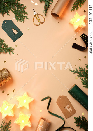 Christmas frame with star candles, fir branches, gift tags, and wrapping supplies on beige background. Copy space for text or design. Christmas frame with star candles, fir branches, gift tags, and wrapping supplies on beige background. Copy space for text or design. 133491315