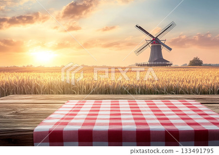 A rustic wooden table with a red-and-white checkered cloth and wheat stalks foregrounds a golden field, a distant windmill, and a beautiful sunset under a dramatic sky. 133491795