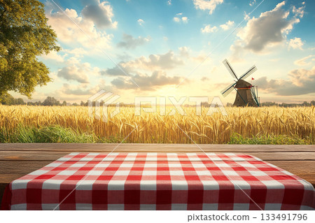 A rustic wooden table with a red-and-white checkered cloth and wheat stalks foregrounds a golden field, a distant windmill, and a beautiful sunset under a dramatic sky. 133491796