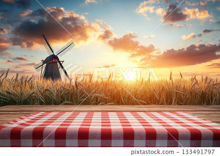 A rustic wooden table with a red-and-white checkered cloth and wheat stalks foregrounds a golden field, a distant windmill, and a beautiful sunset under a dramatic sky. 133491797
