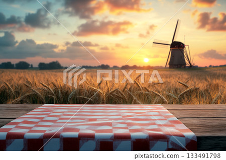A rustic wooden table with a red-and-white checkered cloth and wheat stalks foregrounds a golden field, a distant windmill, and a beautiful sunset under a dramatic sky. 133491798