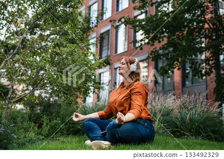 Beautiful mid size woman meditating in park, listening podcast. Beautiful mid size woman meditating in park, listening podcast. 133492329