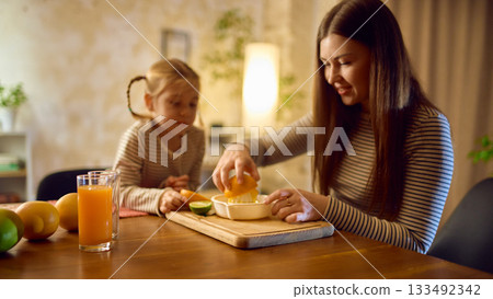 Mother squeezing fresh citrus with daughter observing with orange juice glass standing near 133492342