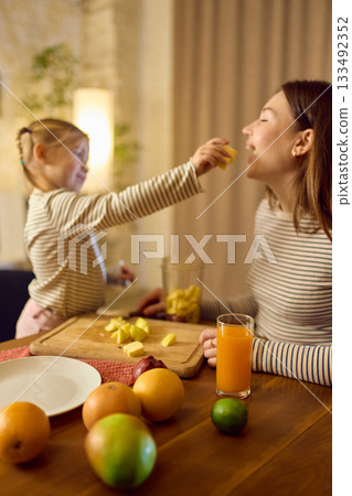 Orange juice on table while daughter feeds fruit to smiling mother in warm playful mood Orange juice on table while daughter feeds fruit to smiling mother in warm playful mood 133492352