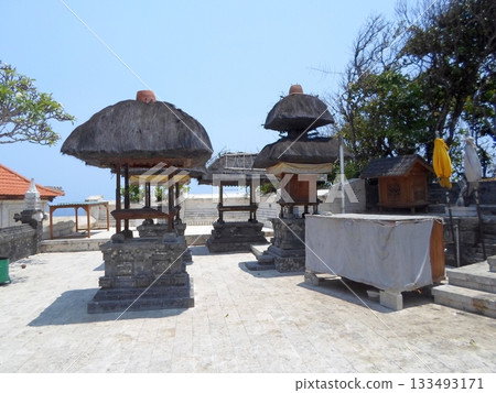 A Temple Courtyard with Traditional Thatched Structures and Stone Elements Under a Bright Sky 133493171