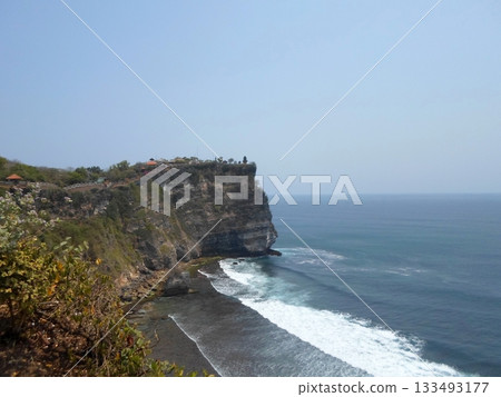 A Cliffside Shoreline with Waves Crashing Against Rocks and Greenery on the Cliff Under a Clear Sky A Cliffside Shoreline with Waves Crashing Against Rocks and Greenery on the Cliff Under a Clear Sky 133493177