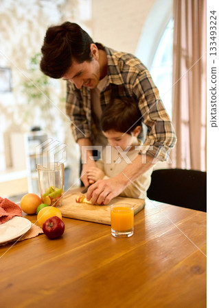 Orange juice in glass on wooden table with father helping son cut fruits in bright mood Orange juice in glass on wooden table with father helping son cut fruits in bright mood 133493224