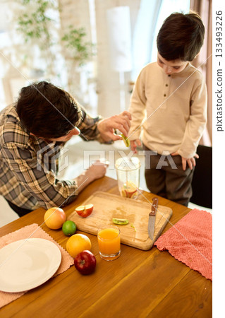 Glass of orange juice with father and son adding fruits to blender in sunny kitchen 133493226