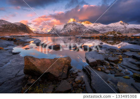 Snowy mountains reflecting on calm lake with stones at sunrise Snowy mountains reflecting on calm lake with stones at sunrise 133495308