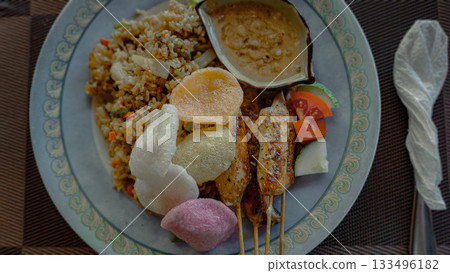Plate of fried rice or nasi goreng and chicken satay skewers served with kropek crackers and peanut sauce on a woven placemat 133496182