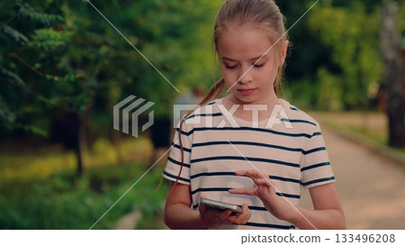 kid smiling while talking on phone outside, kid enjoying phone conversation in park, small child speaking on cell in nature, girl answering call in peaceful park, outdoor mobile chat with little girl 133496208