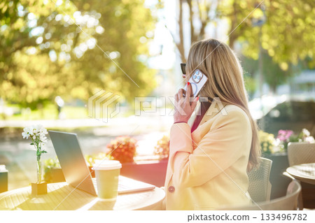 Woman talking on phone while working outdoors at a sunny cafe during the day Woman talking on phone while working outdoors at a sunny cafe during the day 133496782
