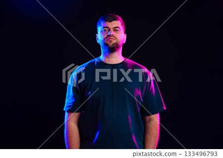 Young man standing confidently against a dark background with colorful lights in the evening Young man standing confidently against a dark background with colorful lights in the evening 133496793