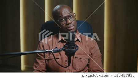 Charismatic Male Host With Glasses Smiling and Looking Directly at Camera While Recording Podcast 133497933