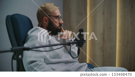 Bearded Podcast Host in Cap and Glasses Gestures While Speaking Into Professional Studio Microphone Bearded Podcast Host in Cap and Glasses Gestures While Speaking Into Professional Studio Microphone 133498470