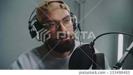 Expressive Bearded Podcast Host Wearing Cap and Glasses Talks Into High Quality Microphone Expressive Bearded Podcast Host Wearing Cap and Glasses Talks Into High Quality Microphone 133498483