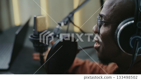 Man Wearing Glasses and Studio Headphones Speaking Energetically Into Pro Microphone Man Wearing Glasses and Studio Headphones Speaking Energetically Into Pro Microphone 133498502
