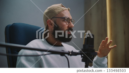 Podcast Host With Beard And Eyewear Explains Point Passionately, Gesturing Beside Microphone Podcast Host With Beard And Eyewear Explains Point Passionately, Gesturing Beside Microphone 133498503