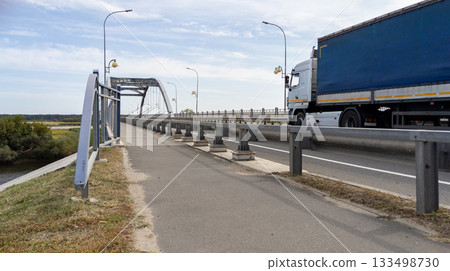 Large blue truck drives along a road across a river bridge against the backdrop of cloudy sky Large blue truck drives along a road across a river bridge against the backdrop of cloudy sky 133498730