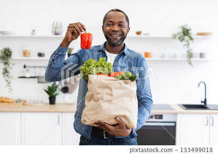 An African American man smiles while holding a bag of fresh groceries in a modern kitchen, including a red bell pepper. 133499018