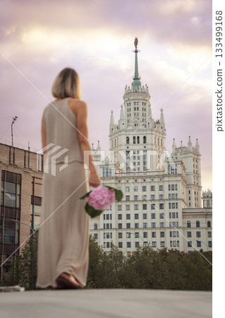 A woman stands at sunset holding a pink hydrangea, looking toward the Kotelnicheskaya Embankment Building rising against a pastel sky A woman stands at sunset holding a pink hydrangea, looking toward the Kotelnicheskaya Embankment Building rising against a pastel sky 133499168