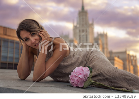 A woman lies on a rooftop with a pink hydrangea beside her, set against the blurred silhouette of the Kotelnicheskaya Embankment Building and warm sky A woman lies on a rooftop with a pink hydrangea beside her, set against the blurred silhouette of the Kotelnicheskaya Embankment Building and warm sky 133499189